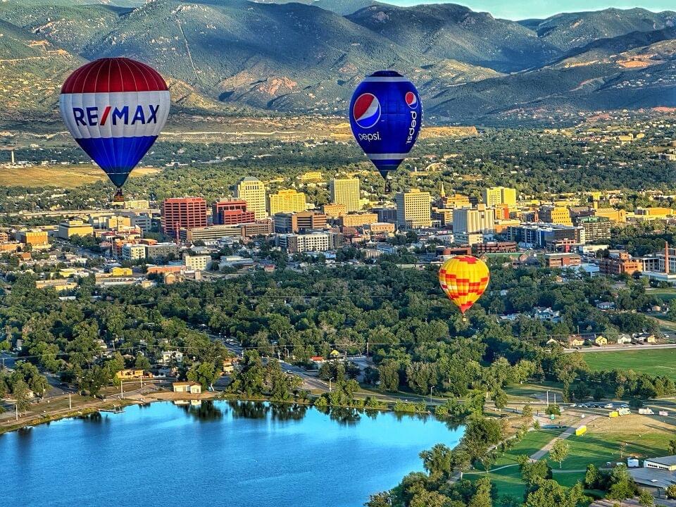 An aerial view of downtown Colorado Springs with three hot air balloons in the sky above Prospect Lake.